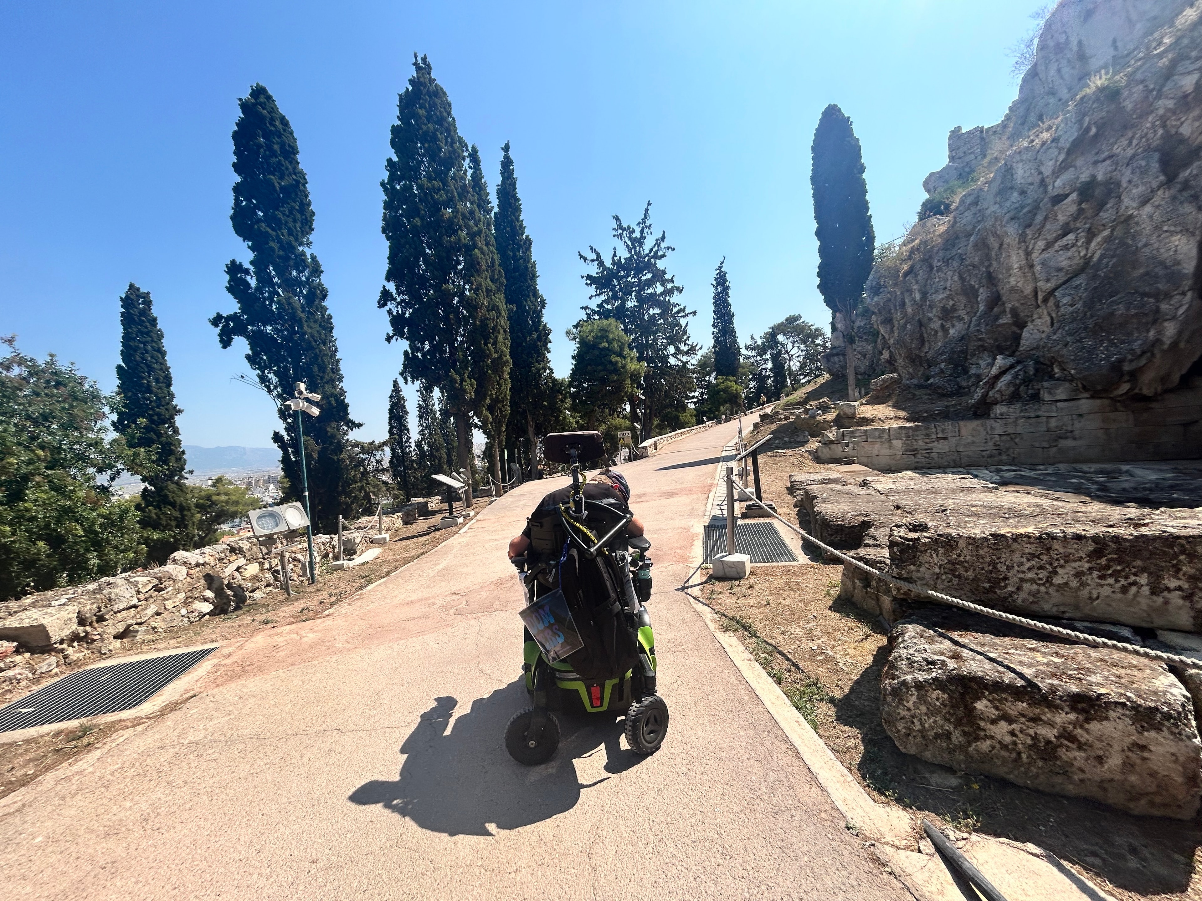 Wide accessible paved path lined with trees leading up toward the Acropolis